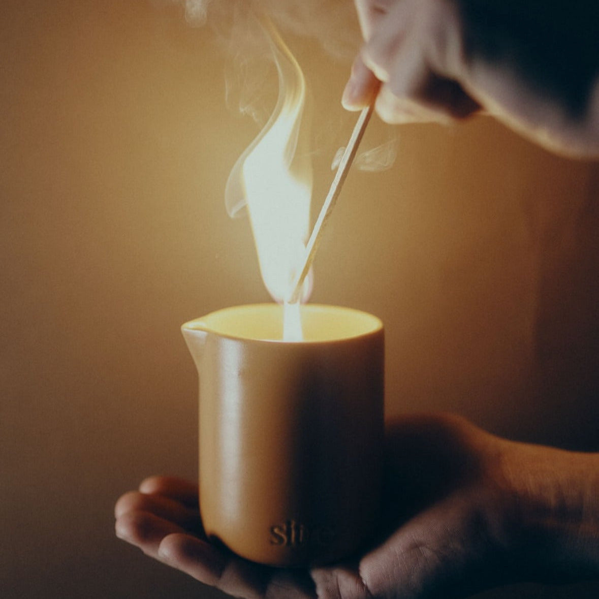 Person lighting a candle with a match, held in hands against a warm background.